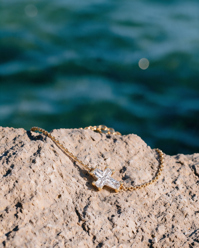 The Eternal Cross Chain Bracelet in 14K recycled yellow gold laid on a rock by the sea.